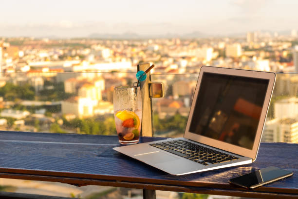 Laptop and glass of summer cocktail on a wooden table, on a bright beautiful panoramic background of the city. mockup Laptop and glass of summer cocktail on a wooden table, on a bright beautiful panoramic background of the city. mockup. Digital Nomad Life: The Best Cities to Work Remotely stock pictures, royalty-free photos & images