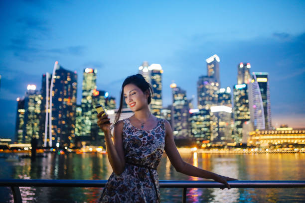 Young fashionable tourist woman in Singapore checking out her smartphone Vintage toned street style portrait of a young fashionable brunette woman, walking in Singapore near the famous bay area. She is wearing a summertime dress, walking among the Singapore skyscrapers, texting on her smartphone, checking apps or maps, figuring where should she go next and do more sightseeing. Singapore: Smart and Safe for travel stock pictures, royalty-free photos & images